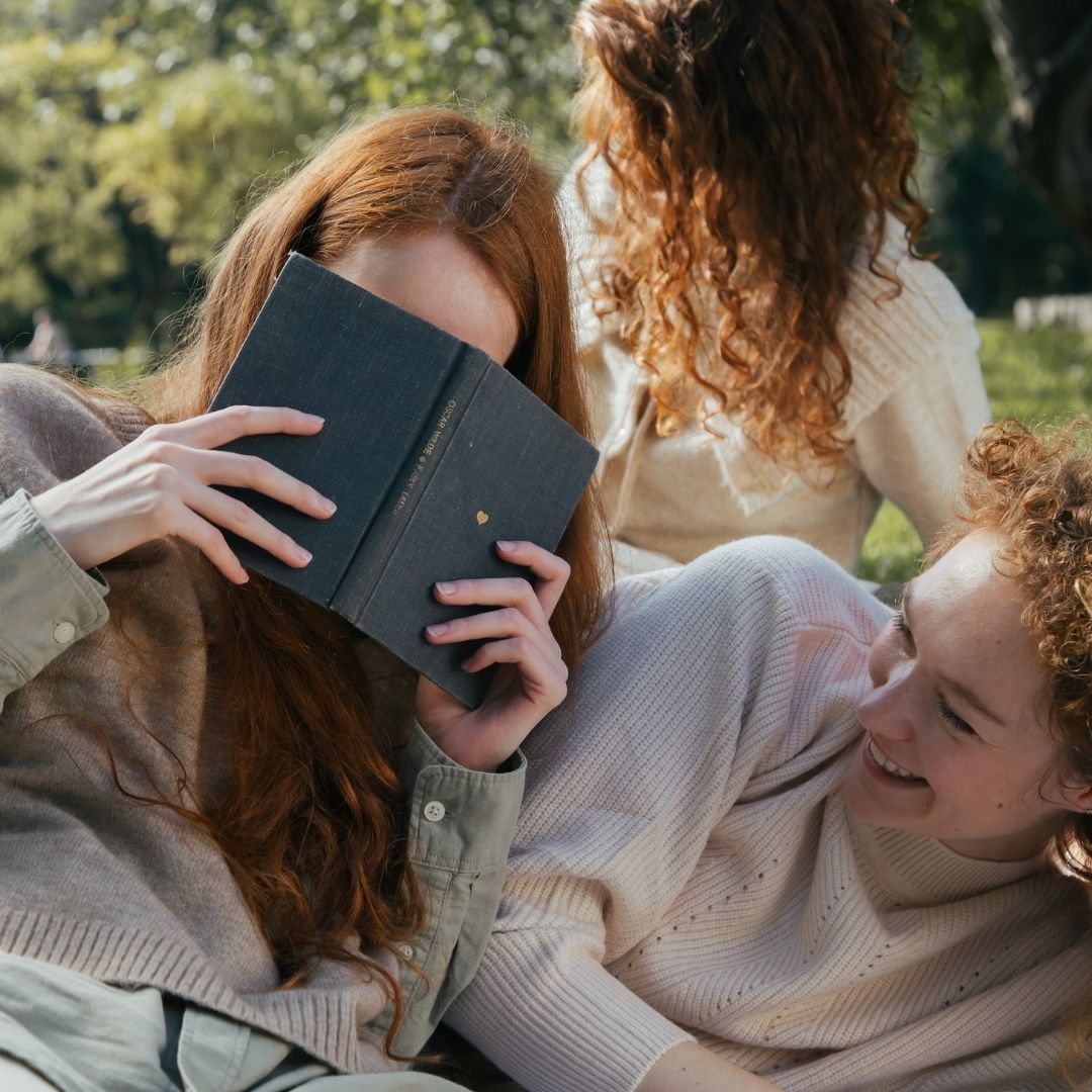 Three women sitting on the grass talking, laughing, and reading.